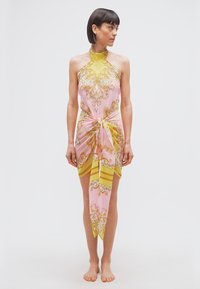 Γυναίκα όρθια, ξυπόλητη, φορούσα φόρεμα με σχέδια σε ροζ και κίτρινο, με κορδέλα στο λαιμό δεμένη στη μέση, κοιτάζοντας προς τα αριστερά της, μπροστά σε απλό φόντο.