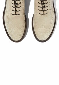 Zapatos brogue de ante beige con diseño de punta de ala, que presentan perforaciones decorativas, cordones marrones y una suela oscura contrastante.