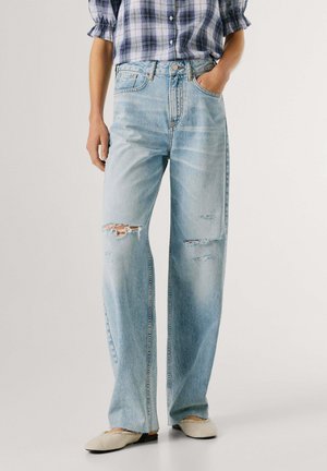 Donna che indossa jeans a gamba dritta strappati a lavaggio chiaro, camicia a quadri e scarpe beige senza lacci, in piedi con una mano in tasca.