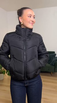 Svart pufferjakke med høy krage og glidelås foran. Har horisontal quilting og sidelommer. Brukes over mørkeblå jeans.