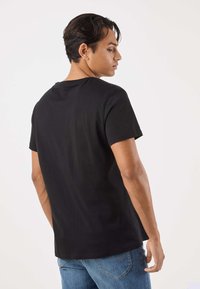 Svart bomullst-shirt med rund halsringning, korta ärmar och en avslappnad passform, sedd bakifrån. Kombineras med blå jeans.