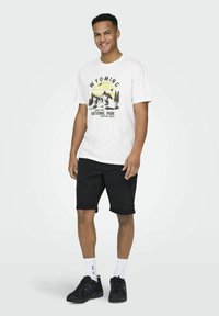 Una camiseta blanca con estampado negro que presenta "WYOMING NATIONAL PARK" y un gráfico de montañas; combinada con pantalones cortos negros enrollados y zapatos negros.