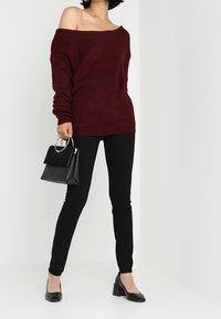 Pull en maille bordeaux à épaules dénudées, jean skinny noir, sac à main noir avec des accents métalliques, et chaussures à talons noires.