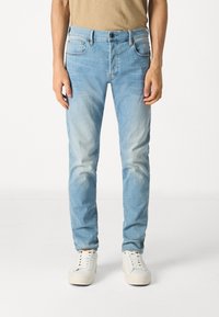 Homme portant un jean bleu clair délavé et des baskets blanches, debout devant un fond uni.