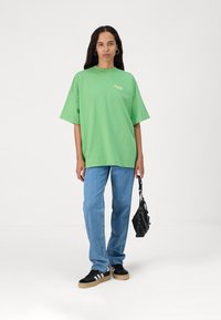Camiseta de algodón verde de gran tamaño con mangas cortas y un pequeño logo amarillo, combinada con jeans de color azul claro y zapatillas negras. Bolsa negra con herrajes.
