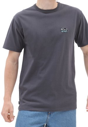 Man wearing dark gray Vans t-shirt and blue jeans, standing with arms relaxed at sides, on a white background.