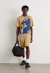 T-shirt graphique beige avec un design bleu, associé à un short beige. La personne porte un sac de sport noir et des chaussures noires avec des chaussettes blanches.