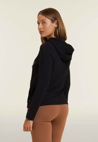 Felpa nera con zip realizzata in tessuto morbido, con cappuccio e tasche frontali, abbinata a leggings marroni. Design semplice con minimi dettagli.