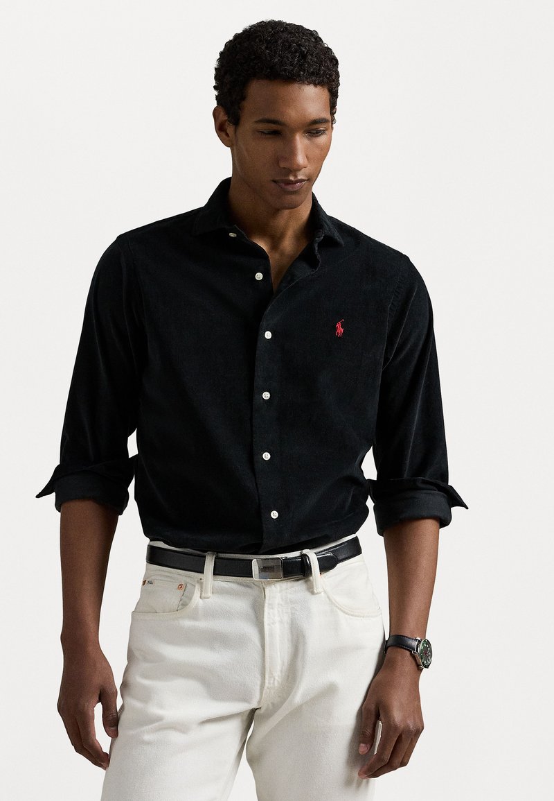 Black button-up shirt made of textured fabric with a small red logo. Paired with light-colored jeans and a black belt featuring a silver buckle.