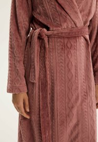 Jasnoróżowy, pluszowy szlafrok z teksturowanym wzorem i geometrycznymi akcentami, z paskiem i długimi rękawami. Miękki, puszysty materiał.