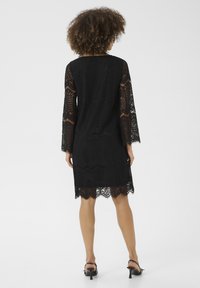 Abito di pizzo nero con maniche lunghe e orlo scallop. Presenta intricati motivi floreali e geometrici, con una silhouette ampia.