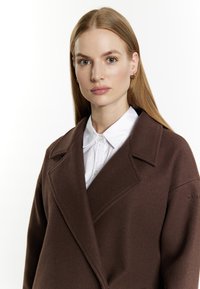 Bruine wollen jas met een grote kraag, voorzien van een ontspannen pasvorm en een voorzak met klep. Witte blouse met kraag eronder. Zachte textuur.