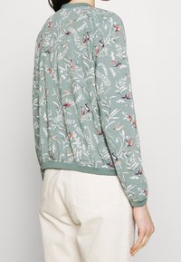 Grön jacka med blommönster och långa ärmar, prydd med fåglar och blad. Mjuk tygkvalitet och rund halsringning med ribbade manschetter och nederkant.