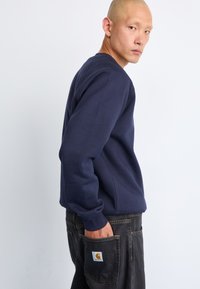 Hombre con sudadera azul marino y jeans oscuros, con la mano en el bolsillo trasero, mirando por encima del hombro contra un fondo blanco liso.