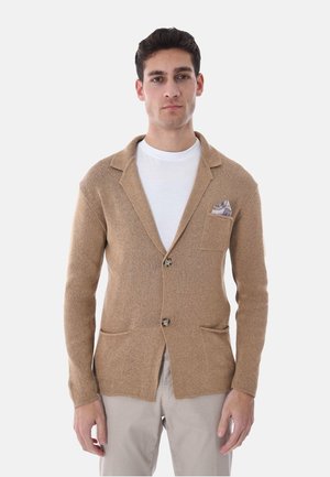 Giovane uomo che indossa un blazer lavorato a maglia color sabbia sopra una camicia bianca e pantaloni beige chiaro, in piedi davanti a uno sfondo chiaro e semplice.