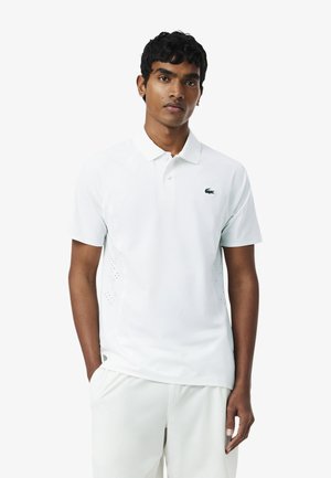 Man draagt een witte poloshirt met korte mouwen en een witte broek, staat met één hand in de zak tegen een effen achtergrond.