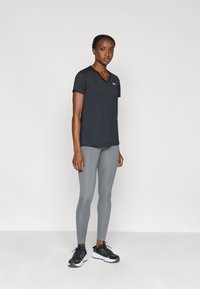 T-shirt de sport noir à col en V associé à des leggings gris. Les chaussures sont noires avec des semelles blanches. Le modèle se tient devant un arrière-plan uni.