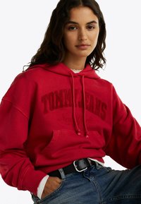 Felpa rossa con la scritta "TOMMY JEANS" e "New York" stampata in rosso scuro. Presenta una tasca a marsupio e cappuccio regolabile con cordino. Indossata con jeans di denim.