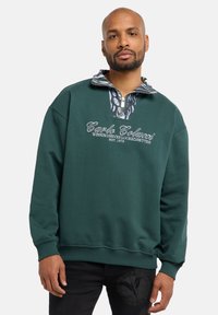 Groene sweater met een hoge kraag, ritssluiting aan de voorkant en een patroon aan de binnenkant van de kraag. Voorzien van geborduurde logo-tekst en geribbelde boorden.