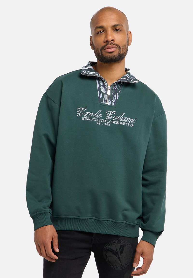 Groene sweater met een hoge kraag, ritssluiting aan de voorkant en een patroon aan de binnenkant van de kraag. Voorzien van geborduurde logo-tekst en geribbelde boorden.