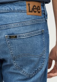 Rücktasche einer Jeans aus Denim mit Lederpatch der Marke Lee und kleinem Stoffetikett auf hellblauem Denim-Stoff, getragen von einer Person.