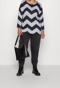 Marineblauw en grijs chevronpatroon lange-mouwen top, gecombineerd met donkergrijze broek en zwart leren veterschoenen. Glad zwart handtas met gestructureerd handvat.