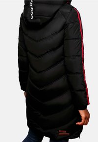 Veste longue noire matelassée avec capuche, rayures rouges sur les manches, logo blanc sur la capuche, portée par une personne vue de dos.