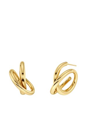 Mujer de piel clara y cabello recogido que lleva pendientes de aro dorados, un collar retorcido dorado, un cuello de tortuga blanco y una prenda de piel beige.