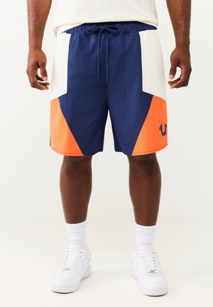 Mann trägt marineblaue, weiße und orangefarbene Shorts mit Kordelzug, weiße Socken und weiße Sneakers und steht vor einem schlichten Hintergrund.
