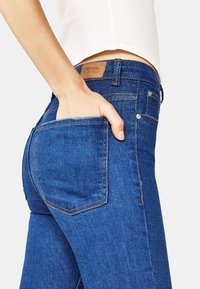 Modré džíny z denimu s těsným střihem, viditelným šitím, zadní kapsou a koženou nášivkou s logem blízko pasu.