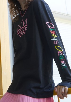Personne portant un t-shirt noir à manches longues avec un motif rose sur la poitrine et le texte "Always Original" en multicolore sur la manche droite.