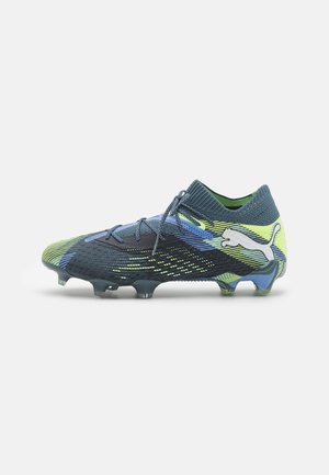 Chaussure de football avec une tige en textile à motif bleu et vert, un col ressemblant à une chaussette, et des crampons en plastique moulé pour une meilleure traction. Logo Puma affiché de manière proéminente.