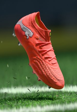 Jasnopomarańczowy but piłkarski z teksturowaną cholewką, stożkowatym kształtem i srebrnym logo Puma; uniesiony nad zieloną trawą, z unoszącymi się cząstkami darni.