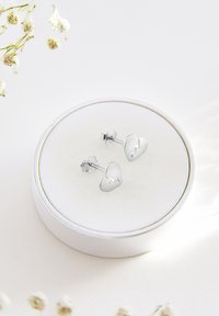 Silver hjärtformade örhängen med en trasig hjärtdesign, utställda på en rund vit kudde inuti en cirkulär låda. Minimalistisk stil.