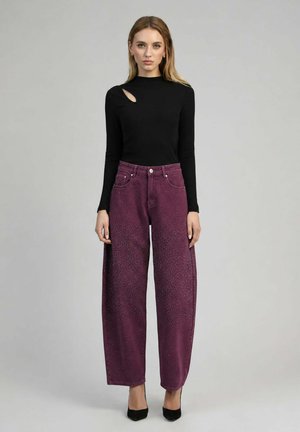 Femme portant un haut noir à manches longues avec une découpe à l'épaule et un pantalon violet taille haute à jambes larges avec de petits motifs à pois.