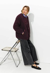 Veste en polaire bordeaux avec un col rond et des boutons, assortie à un jean noir ample et des ballerines noires à clous. Chemise bleu clair en dessous.