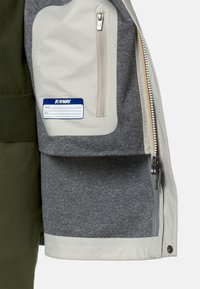 Giacca leggera con interno grigio e esterno color crema. Presenta una tasca con zip e un'etichetta per nome e email.