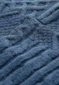 Gros plan d’un tissu bleu doux et texturé avec un motif torsadé en relief, montrant des fibres de laine détaillées et des variations de couture.