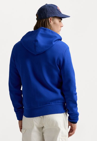 Jovem vestido com um hoodie azul e um boné azul-marinho, visto de costas, com calças de cor clara contra um fundo simples.