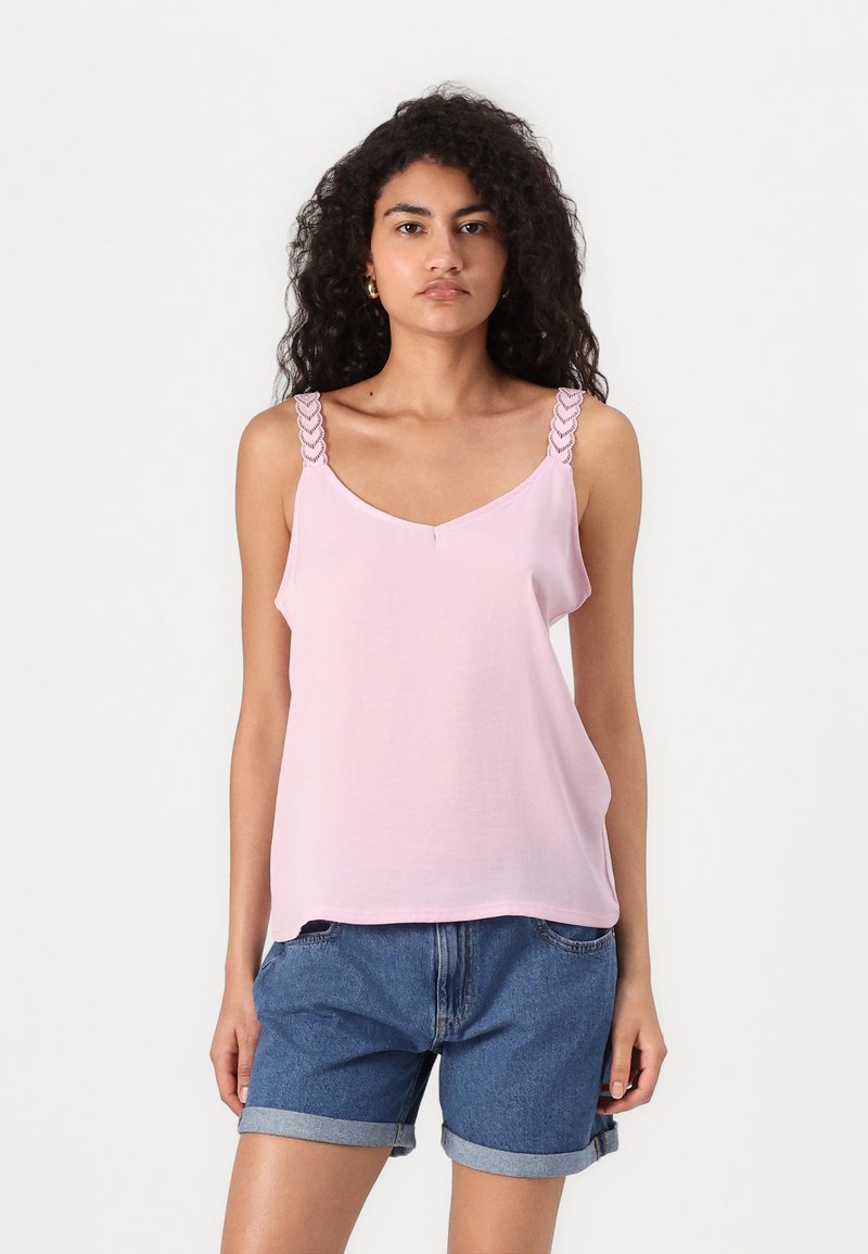 Haut sans manches rose clair avec des bretelles tressées, décolleté en V, tissu doux et coupe décontractée. Porté avec des shorts en denim retroussés à l'ourlet.