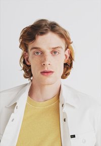 Chaqueta de mezclilla blanca sobre una camiseta amarilla. El sujeto tiene el cabello rizado castaño claro y pecas prominentes, con ojos verdes en una expresión neutral.