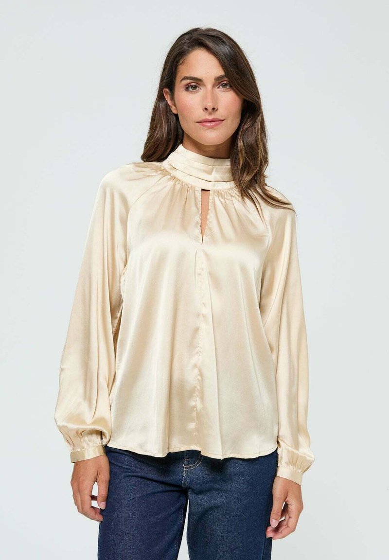 Blouse en satin crème à col haut, avec un détail en trou de serrure et des manches rondes, présentant une texture douce et une coupe décontractée, associée à un jean sombre.