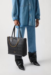 Borsa tote nera in pelle goffrata, con una fascia in catena dorata, doppi manici e logo. Il modello indossa una camicia di jeans e jeans blu.