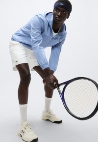 Casaco azul claro com fecho éclair, calções brancos, meias brancas e sapatos de ténis bege. A segurar uma raqueta de ténis com pega texturada e cordas estampadas.