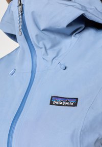 Ανοιχτό μπλε αδιάβροχο μπουφάν με πλήρη φερμουάρ, κουκούλα και λογότυπο της Patagonia στο στήθος, με αντίθετη ραφή και λεπτομέρειες.