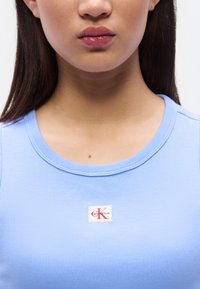 Jeune femme portant un haut sans manches côtelé bleu clair avec un patch logo Calvin Klein sur la poitrine, montrée du menton à la partie supérieure du torse.
