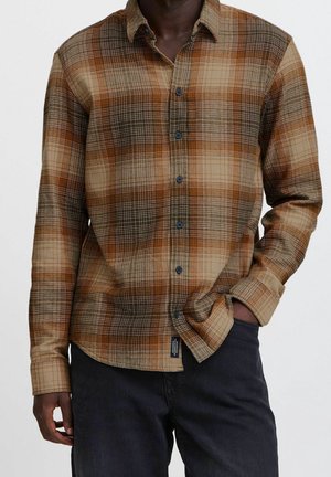 Muž nosící hnědou, béžovou a oranžovou károvanou košili na knoflíky s dlouhým rukávem a tmavé kalhoty, ruka v kapse, na jednoduchém pozadí.