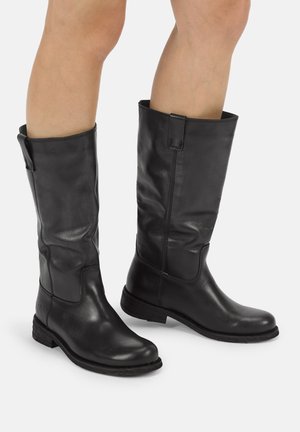 Person, die schwarze Lederstiefel bis zur Mitte der Waden mit niedrigen Absätzen und abgerundeten Zehen trägt, steht vor einem einfachen hellen Hintergrund.