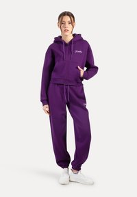 Lilla cropped hoodie med frontlynlås og kængurulomme, parret med matchende tapered joggers og hvide sneakers. Glat stoftekstur.