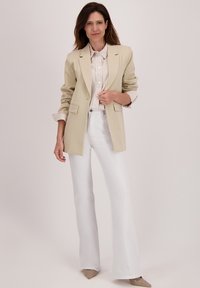 Beige blazer met twee voorkeurszakken, gecombineerd met een lichtgestreept shirt, witte flared broek en beige puntige schoenen.